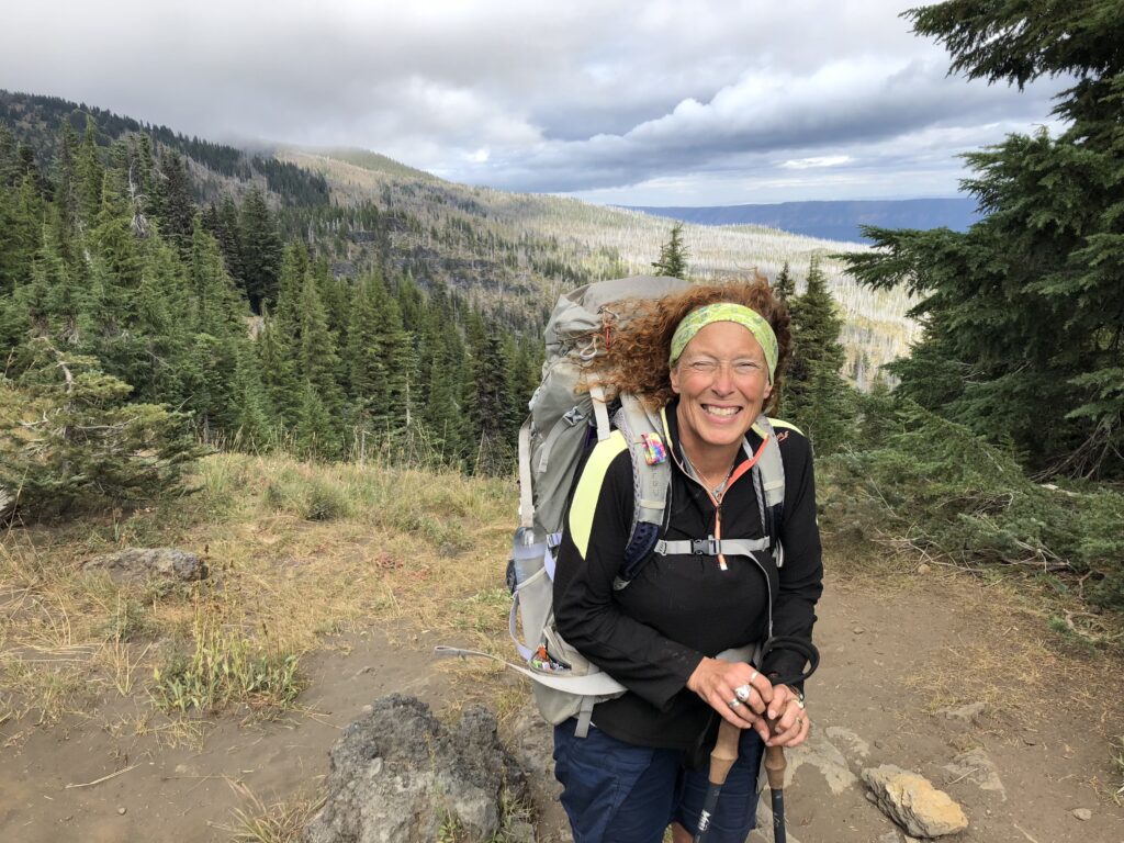 Roberta King, cannabis PR professional poses on the Pacific Crest Trail. She has red curly hair with trekking poles and a gray backpack with Oregon Mountains in the background.